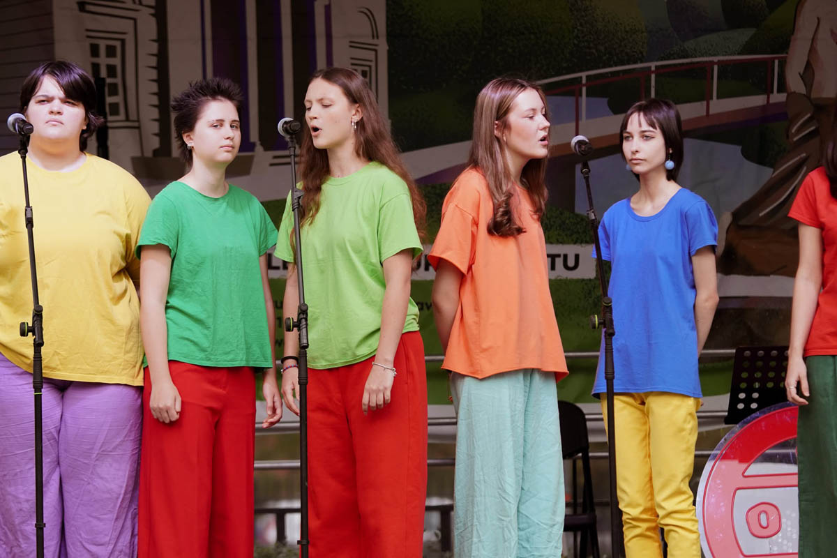 Grupa uczestniczek Teatru NORA ZSŁ w kolorowych koszulkach ustawiona przy mikrofonach na scenie.