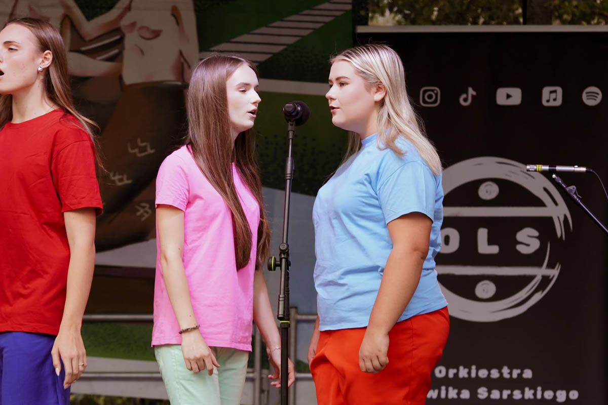 Trzy uczestniczki Teatru NORA ZSŁ w czerwonej, różowej i niebieskiej koszulce podczas występu na scenie.