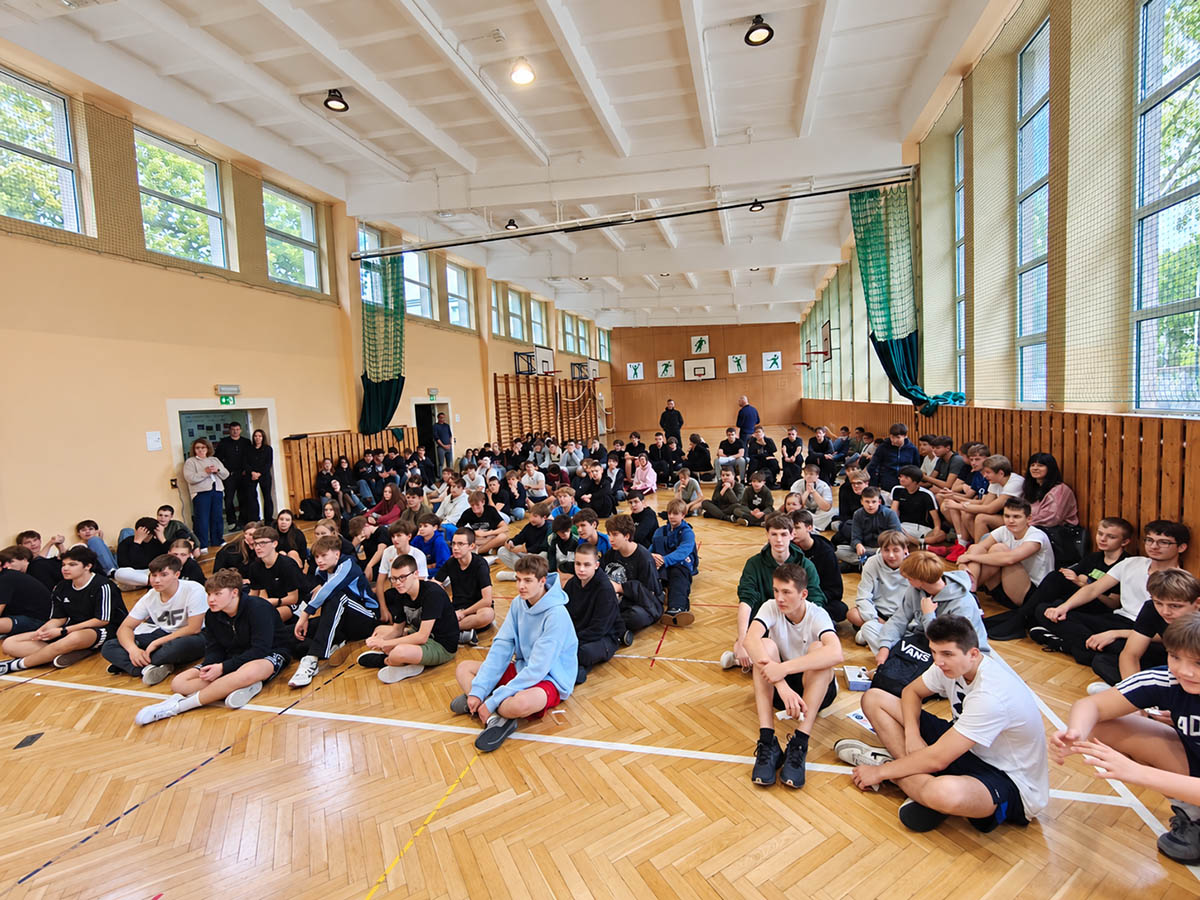 Uczniowie klas pierwszych siedzący na parkiecie hali sportowej i kibicujący podczas Dnia Sportu Szkolnego Pierwszaki na start
