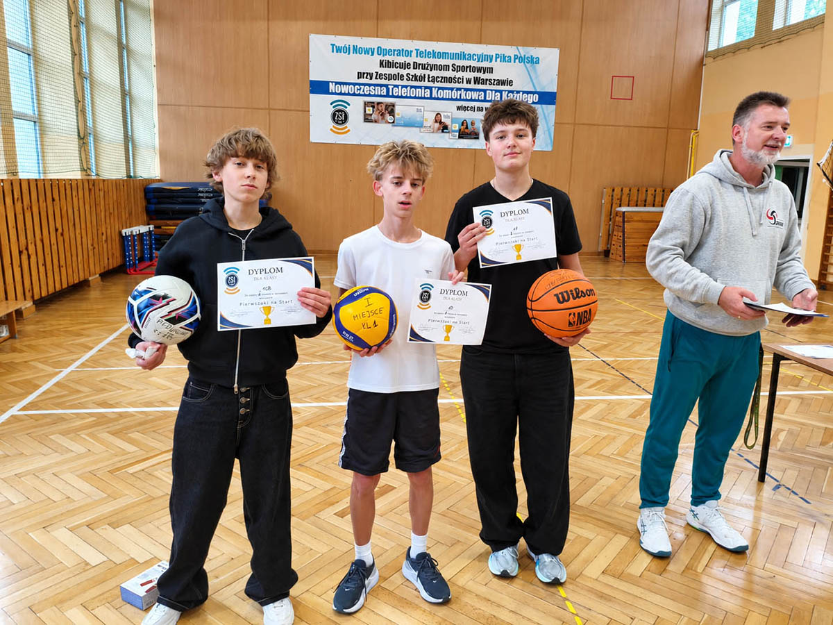 Trzej uczniowie na hali sportowej prezentujący zdobyte dyplomy oraz piłki za zwycięstwo w zawodach Pierwszaki na start