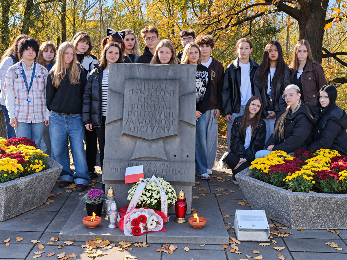 Uczniowie ZSŁ stoją przy miejscu pamięci udekorowanym kwiatami, zniczami i biało-czerwoną wiązanką