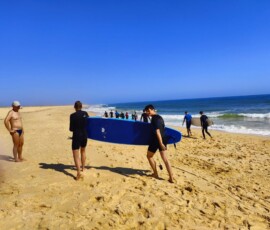 Dwóch uczniów niesie dużą niebieską deskę podczas zajęć surfingowych.