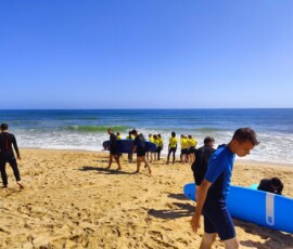 Uczeń niesie niebieską deskę surfingową podczas zajęć na Praia de Faro.