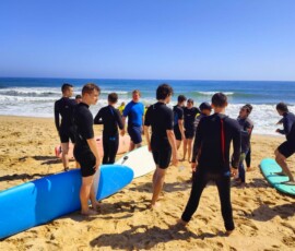 Uczestnicy niosą deski surfingowe po plaży przed ćwiczeniami.