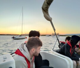 Uczestnicy mobilności siedzą w kamizelkach ratunkowych na pokładzie łodzi podczas rejsu o zachodzie słońca.