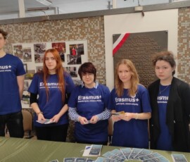 Uczniowie ZSŁ w koszulkach projektu stoją przy stoisku promującym Erasmus+ podczas wydarzenia szkolnego.