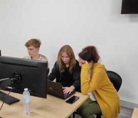 Uczniowie pracują przy monitorach, a prowadząca pomaga im podczas zajęć.