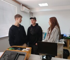 Troje uczniów stoi przy stole z urządzeniami podczas zajęć praktycznych.