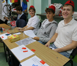 Uczniowie w czerwonych nakryciach głowy przy stoisku udekorowanym flagą Turcji.