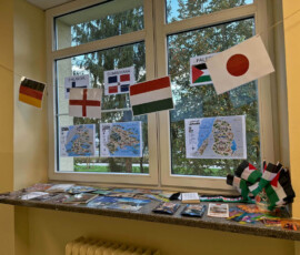 Flagi kilku państw zawieszone nad mapami i materiałami edukacyjnymi na szkolnym oknie.